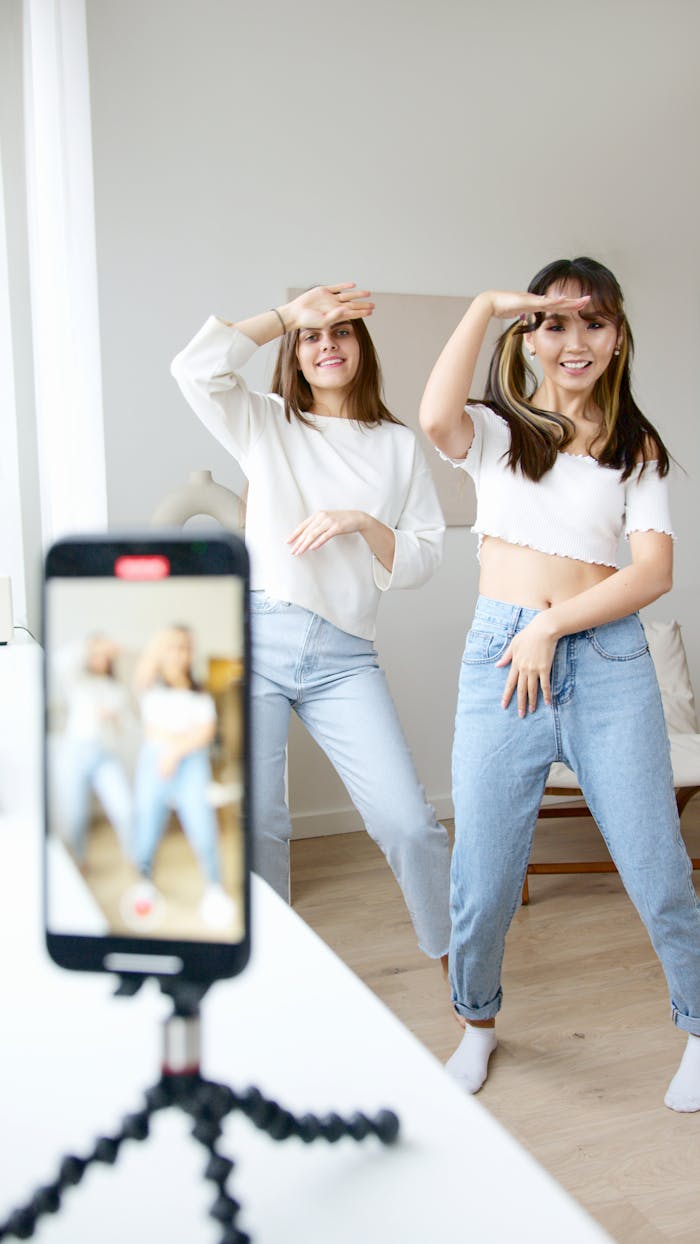 Two young women dancing and recording with a smartphone, wearing casual outfits indoors.